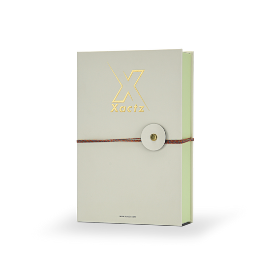Luxury cream book style perfume gift box three quarter angle view showing embossed gold foil Xactz logo soft-touch matte lamination pale green interior spine and multicolour rope closure with gold metal rivet