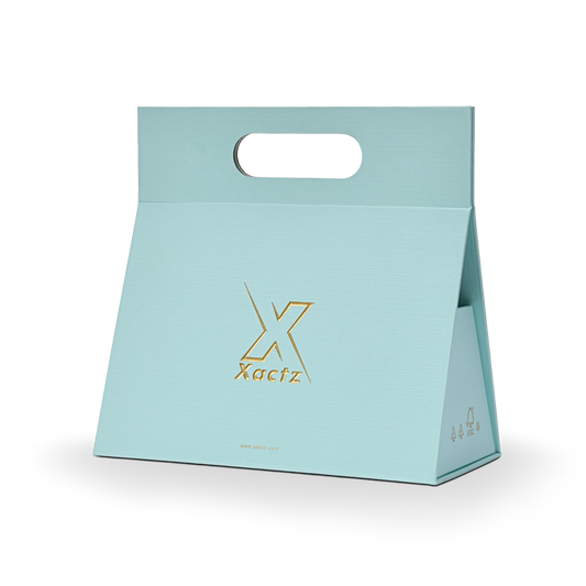 Three quarter angled view of a custom foldable gift box bag with handle in sky blue linen specialty paper showing the tapered trapezoidal body shape with the embossed matte gold foil Xactz logo on the front panel and FSC certification marks in gold on the side panel