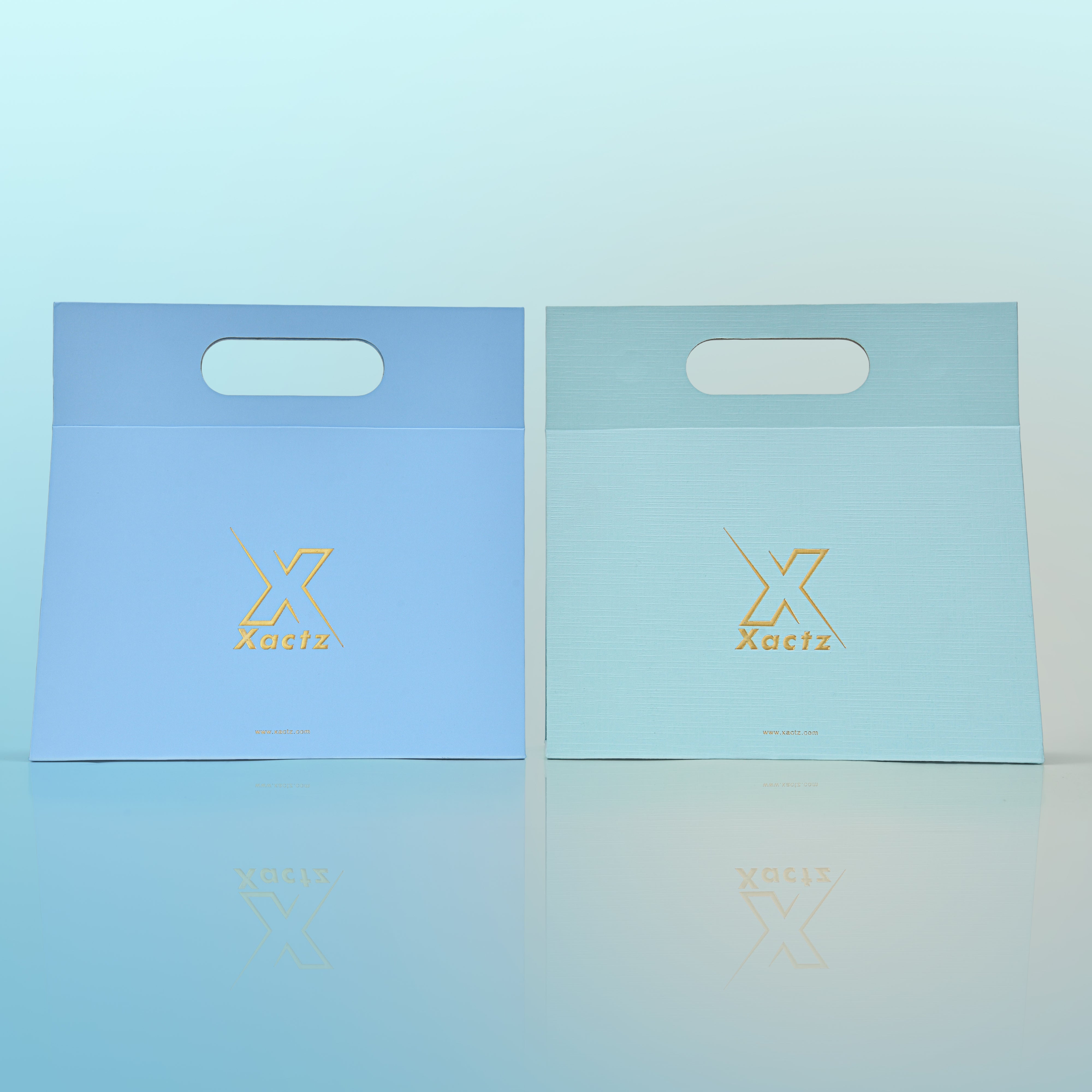 Front view of two custom foldable gift box bags with handle side by side showing the sky blue art paper version on the left and the mint green textured specialty paper version on the right both featuring embossed gold foil Xactz logos