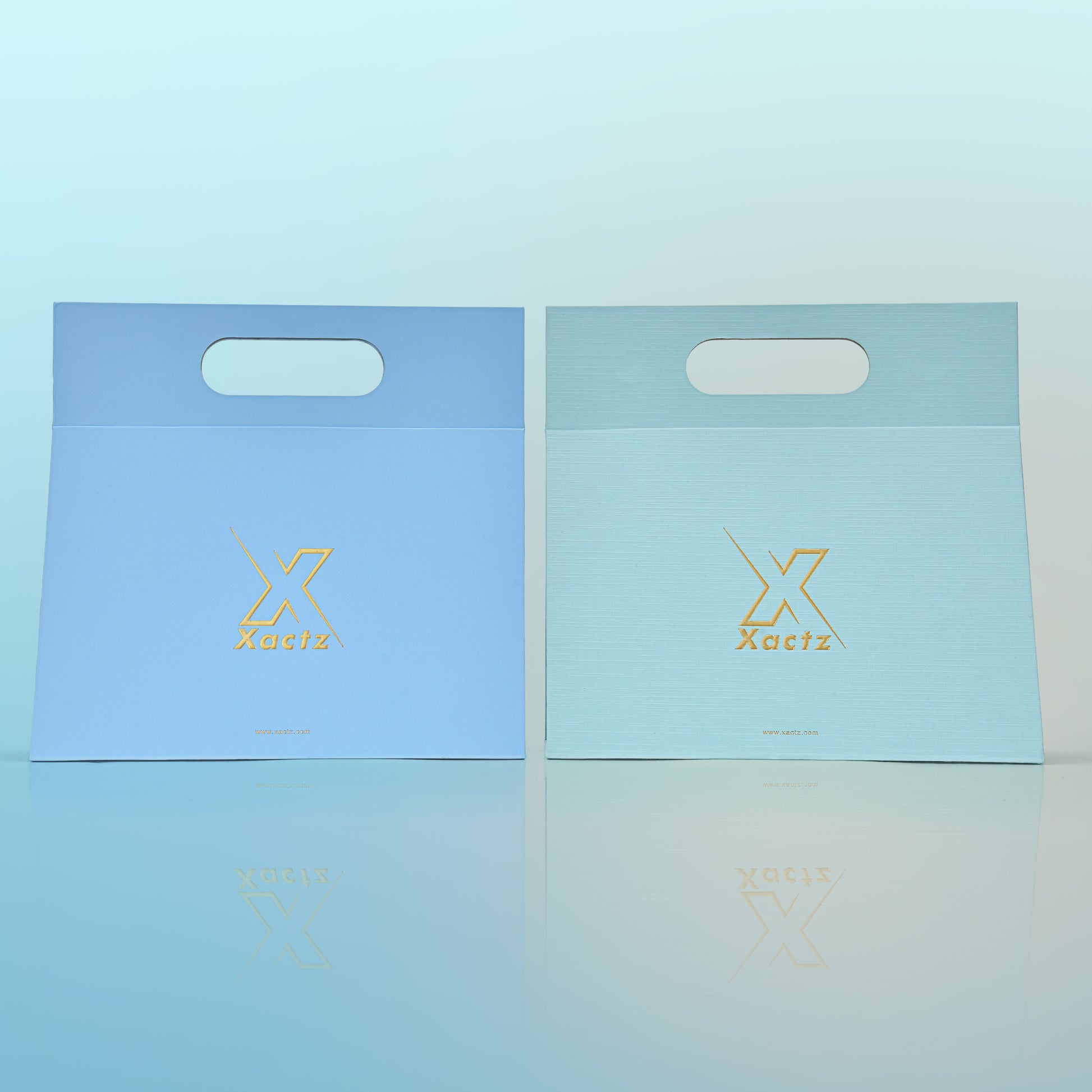 Front view of two custom foldable gift box bags with handle side by side showing the sky blue art paper version on the left and the mint green textured specialty paper version on the right both featuring embossed gold foil Xactz logos