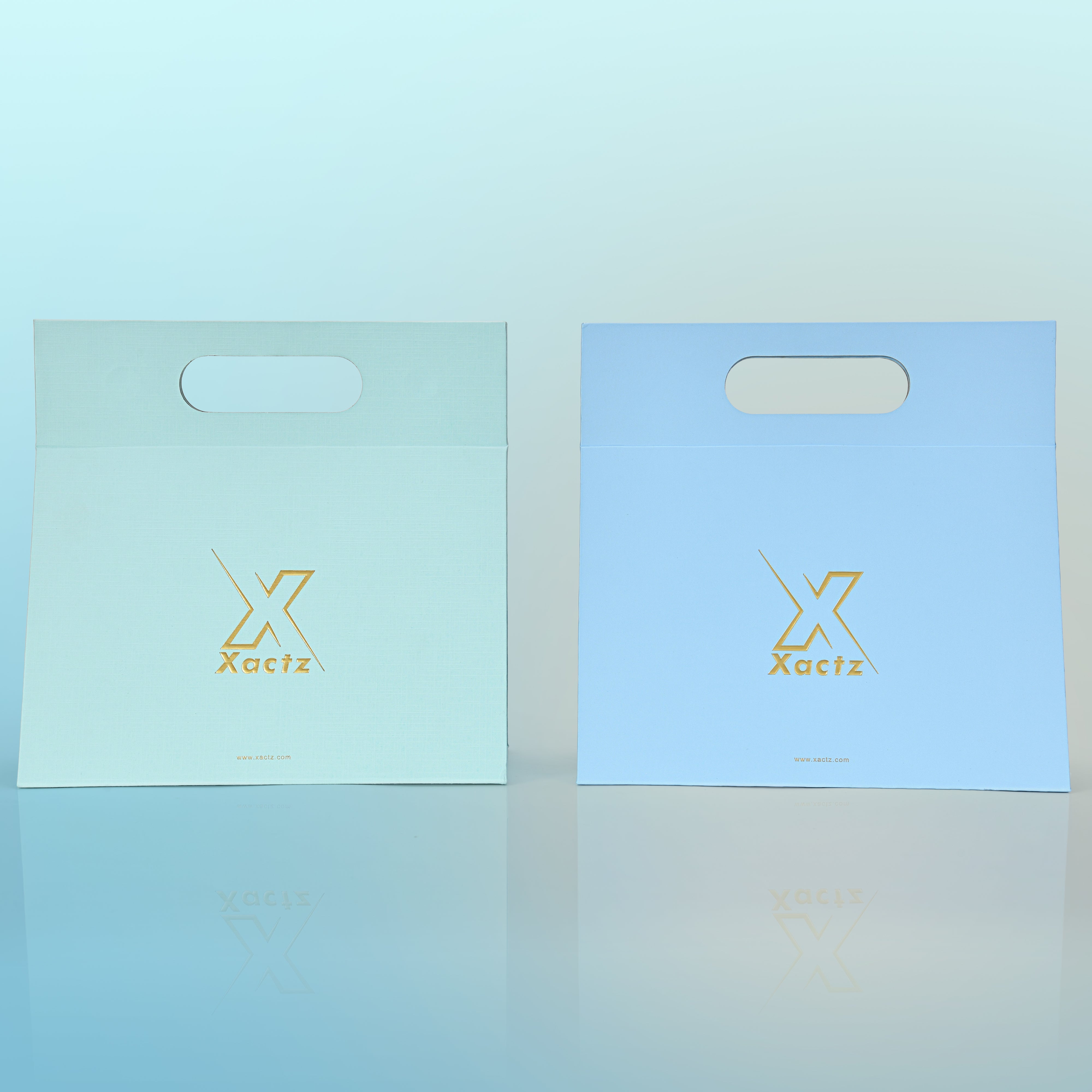 Front view comparison of two custom foldable gift box bags with handle showing the sky blue linen specialty paper version with embossed matte gold foil Xactz logo on the left and the powder blue linen specialty paper version with the same embossed matte gold foil logo on the right both with die cut carry handle cutouts