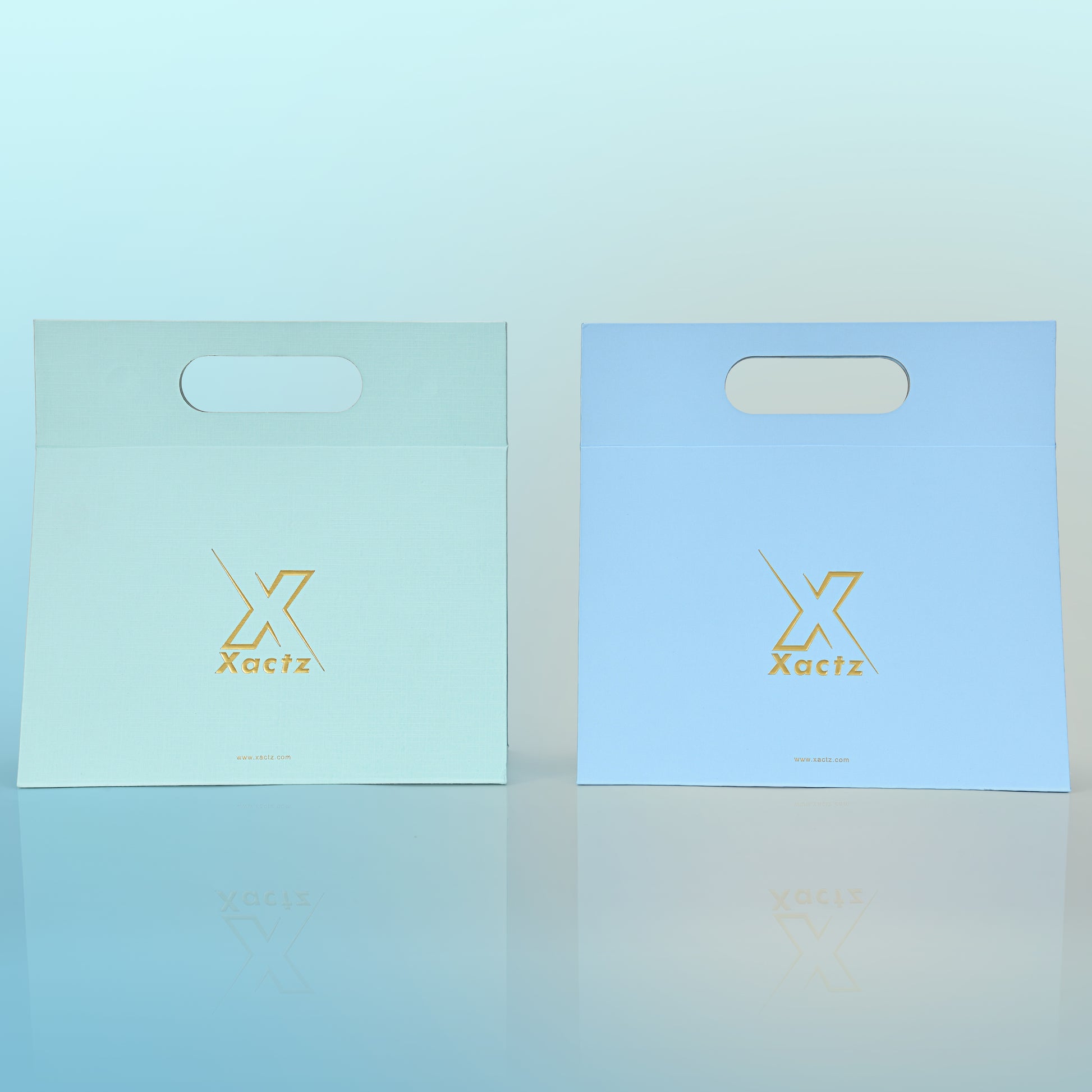 Front view comparison of two custom foldable gift box bags with handle showing the sky blue linen specialty paper version with embossed matte gold foil Xactz logo on the left and the powder blue linen specialty paper version with the same embossed matte gold foil logo on the right both with die cut carry handle cutouts