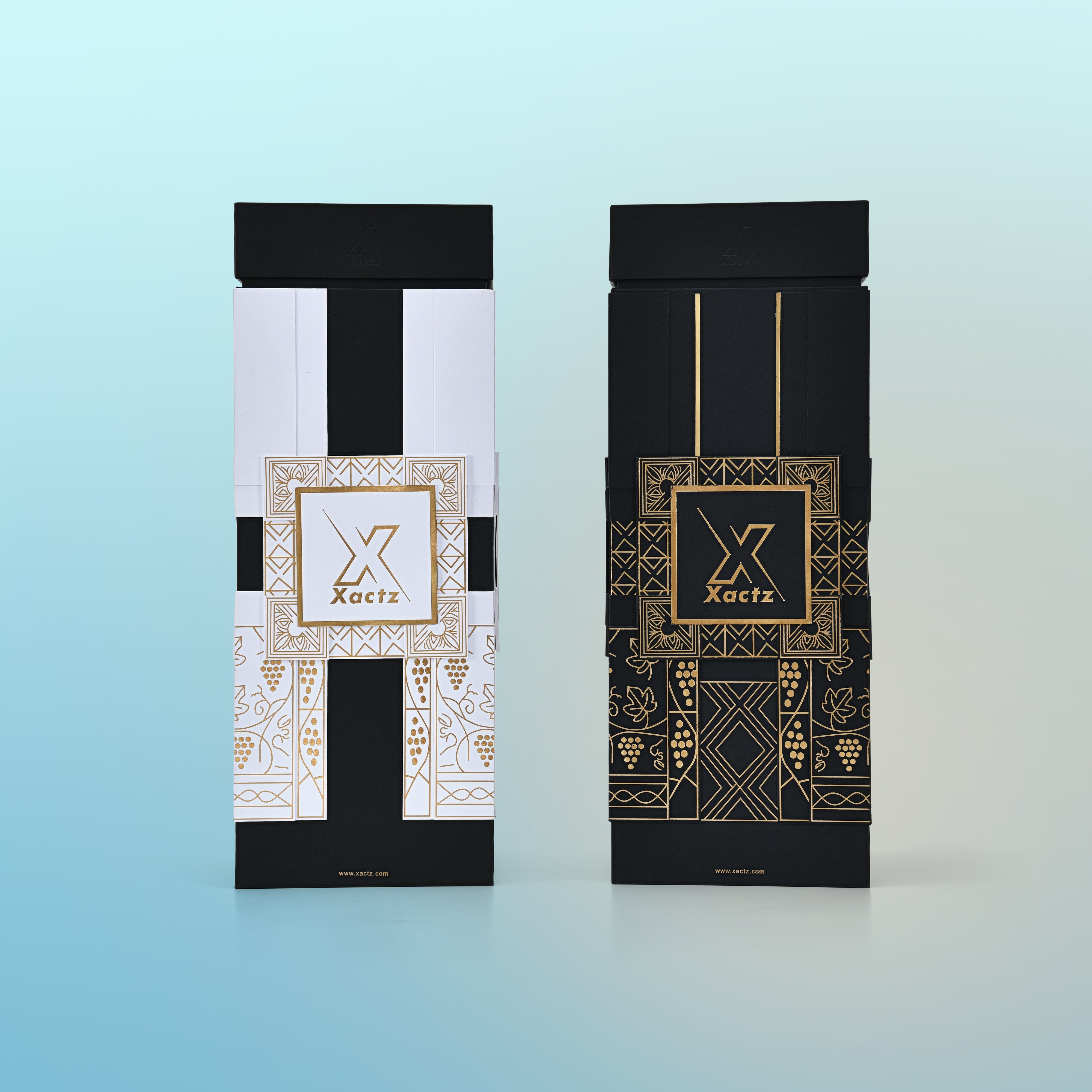 Three quarter angled view of two assembled custom black rigid wine gift boxes showing the white body gold foil version and the all black body gold foil version side by side both with matte black collared lids and full gold foil geometric grape vine illustrations wrapping all four panels