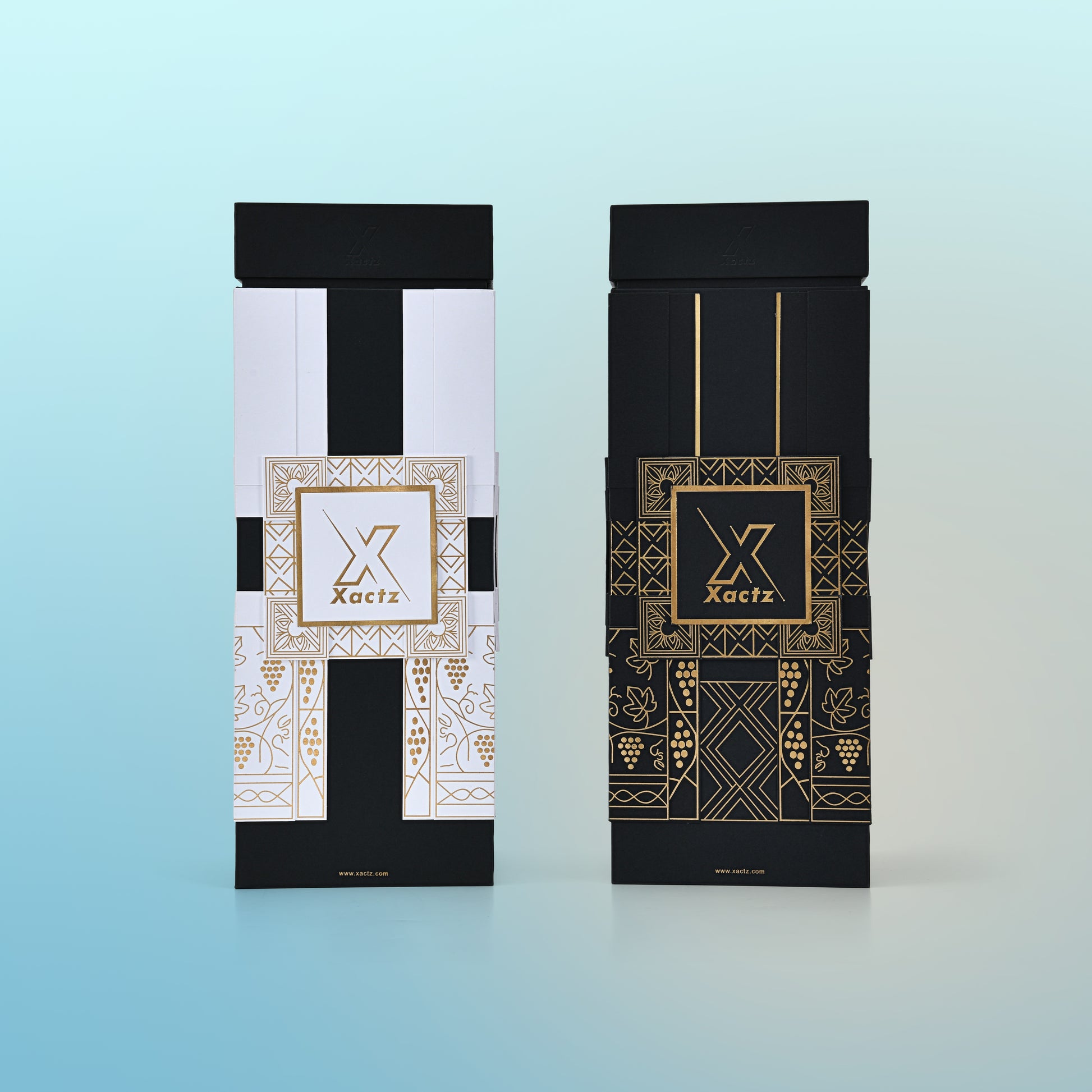 Three quarter angled view of two assembled custom black rigid wine gift boxes showing the white body gold foil version and the all black body gold foil version side by side both with matte black collared lids and full gold foil geometric grape vine illustrations wrapping all four panels