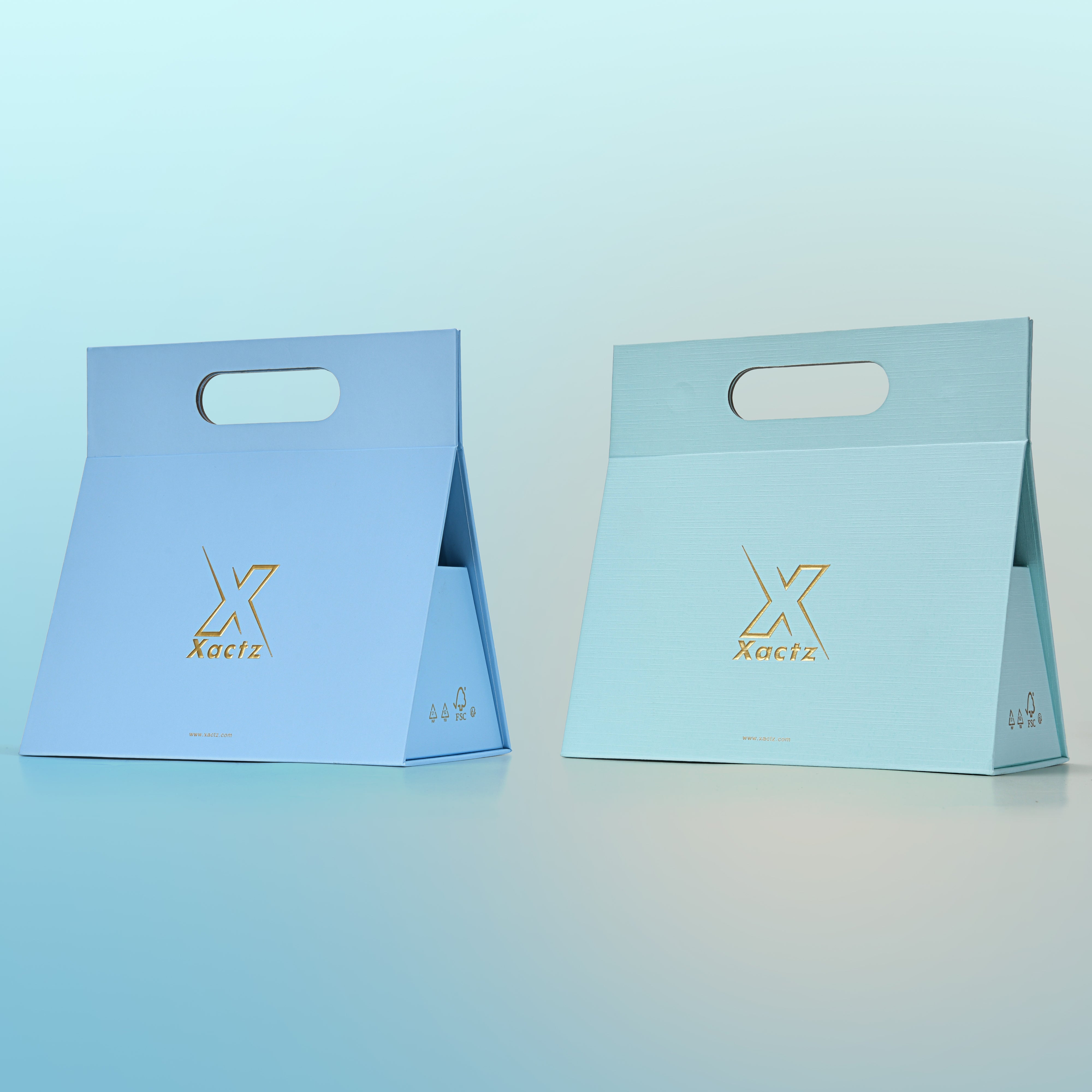 Three quarter angled view of two assembled custom foldable gift box bags with handle showing the sky blue smooth art paper version and the mint green textured specialty paper version both with gold foil logos and FSC certification marks
