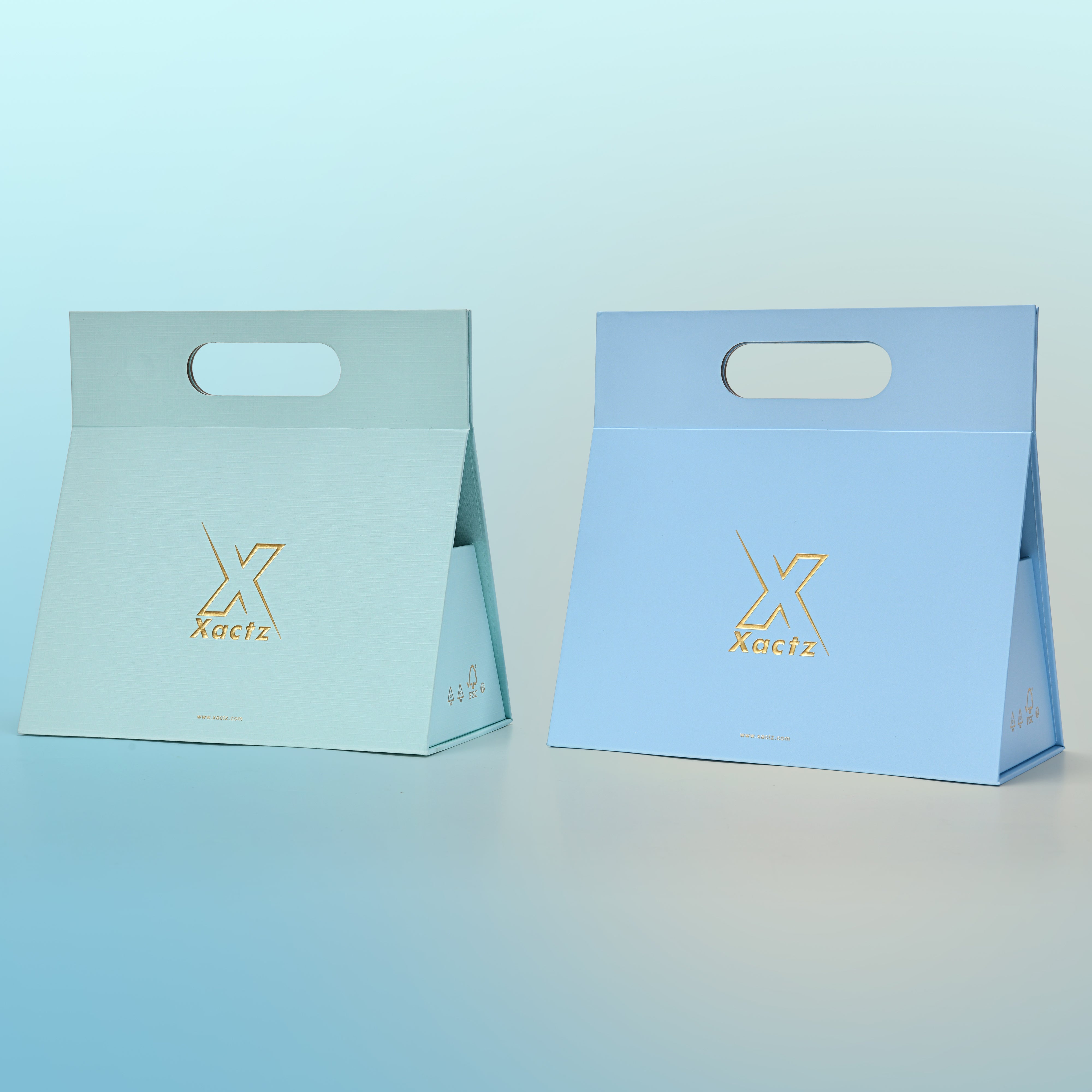 Three quarter angled view of two assembled custom foldable gift box bags with handle showing the sky blue linen specialty paper version and the powder blue linen specialty paper version side by side both displaying the tapered trapezoidal body shape the embossed matte gold foil Xactz logo and the die cut carry handle at the top
