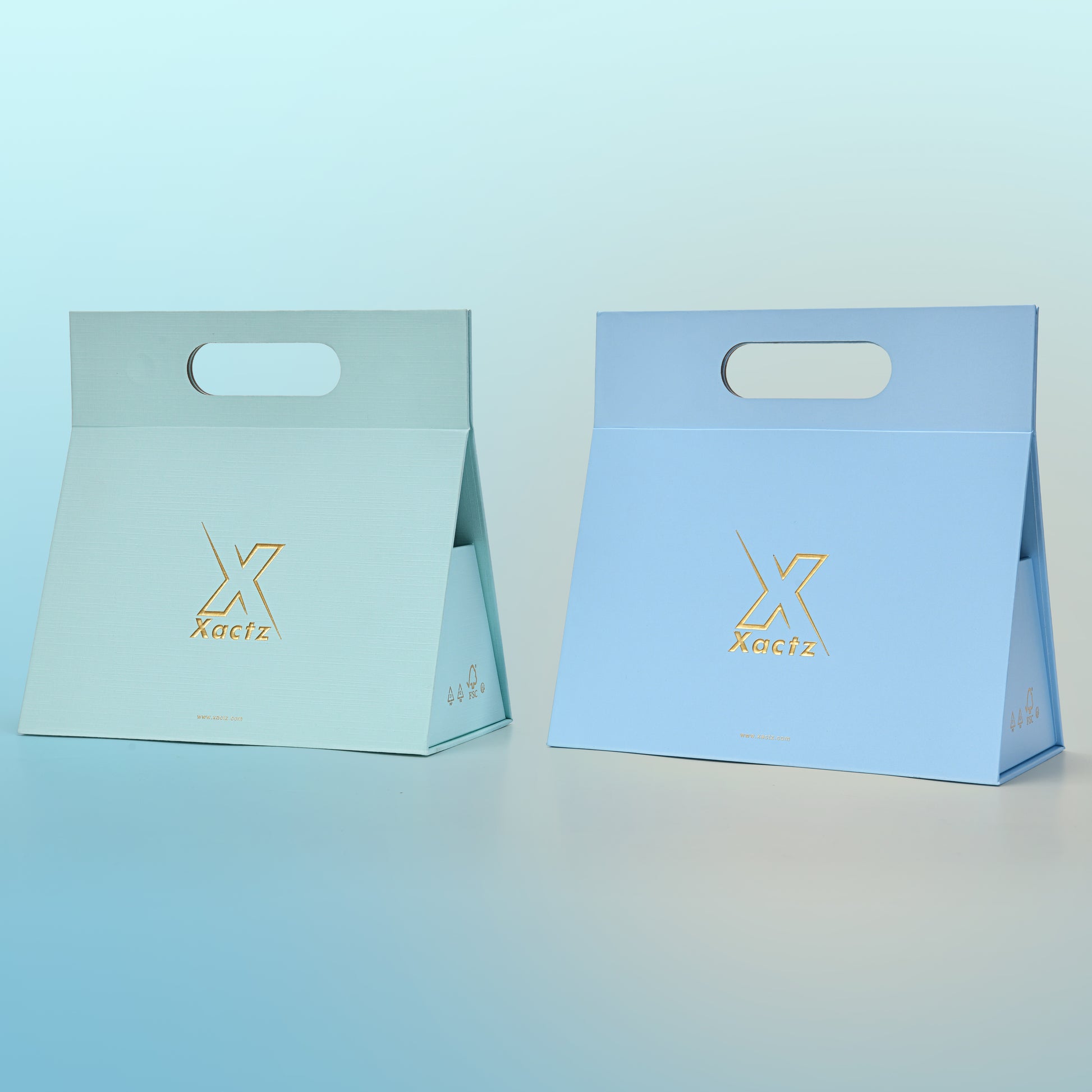 Three quarter angled view of two assembled custom foldable gift box bags with handle showing the sky blue linen specialty paper version and the powder blue linen specialty paper version side by side both displaying the tapered trapezoidal body shape the embossed matte gold foil Xactz logo and the die cut carry handle at the top