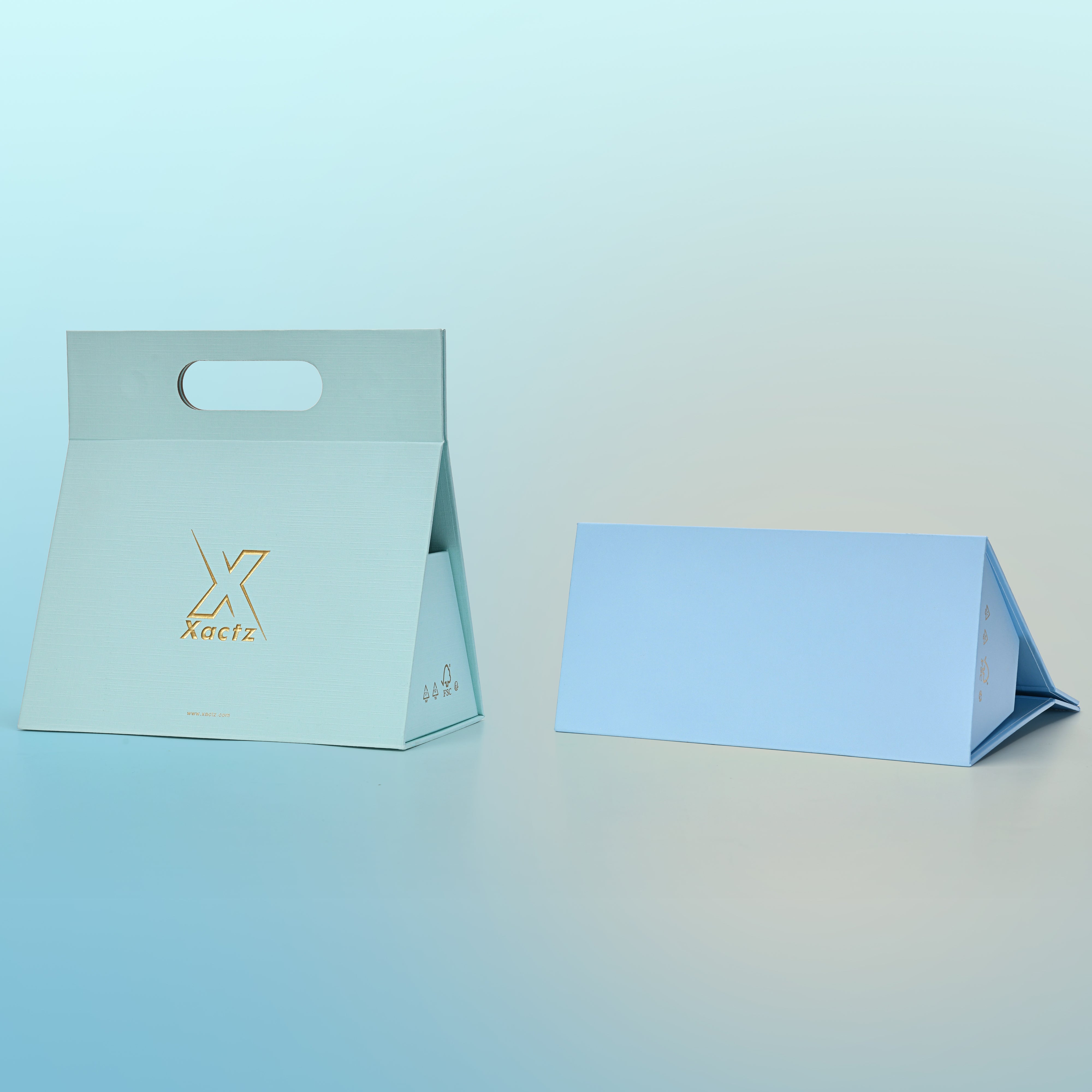 Lifestyle view of two custom foldable gift box bags with handle showing the sky blue linen specialty paper version fully assembled and upright with the embossed matte gold foil Xactz logo on the front panel and the powder blue linen specialty paper version folded flat beside it displaying the flat pack storage format of the foldable gift box bag construction