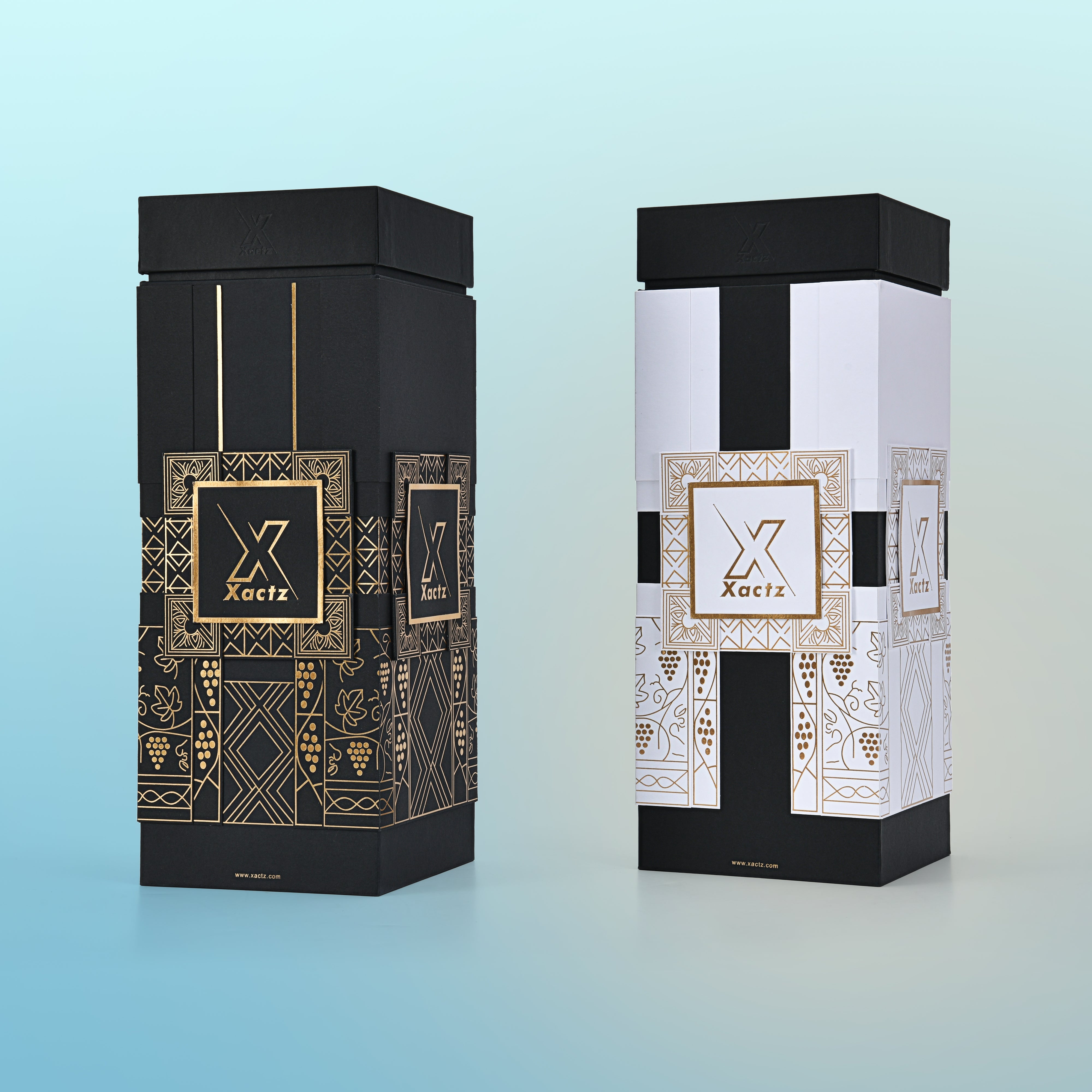 Three quarter angled view of two assembled custom rigid wine gift boxes showing the all black body gold foil version and the white and black body gold foil version side by side both with matte black collared lids and full gold foil geometric grape vine illustrations wrapping all four panels