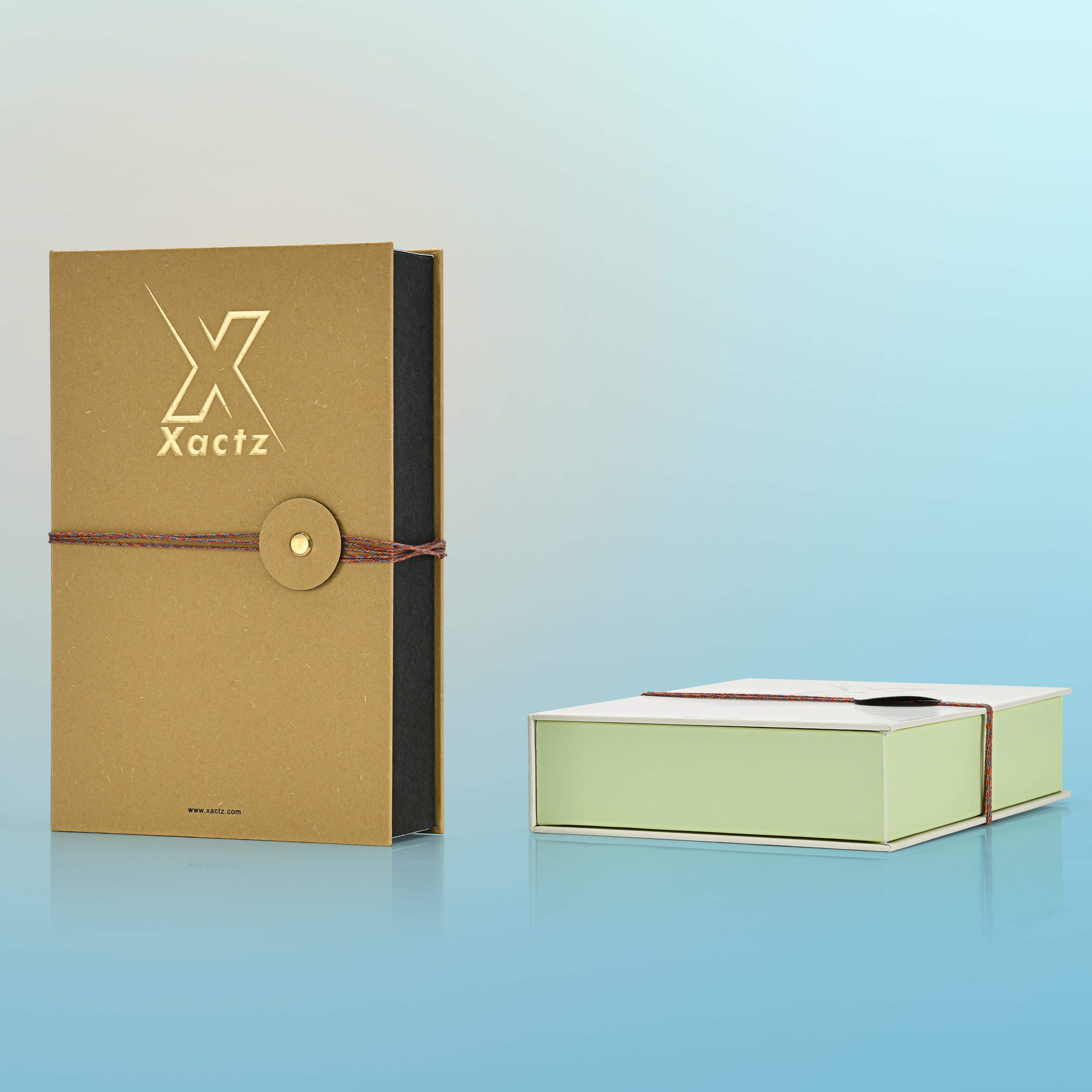 Product display showing a standing ochre yellow custom rigid book style perfume box next to a closed light grey and light green book style box lying flat on a reflective light blue background