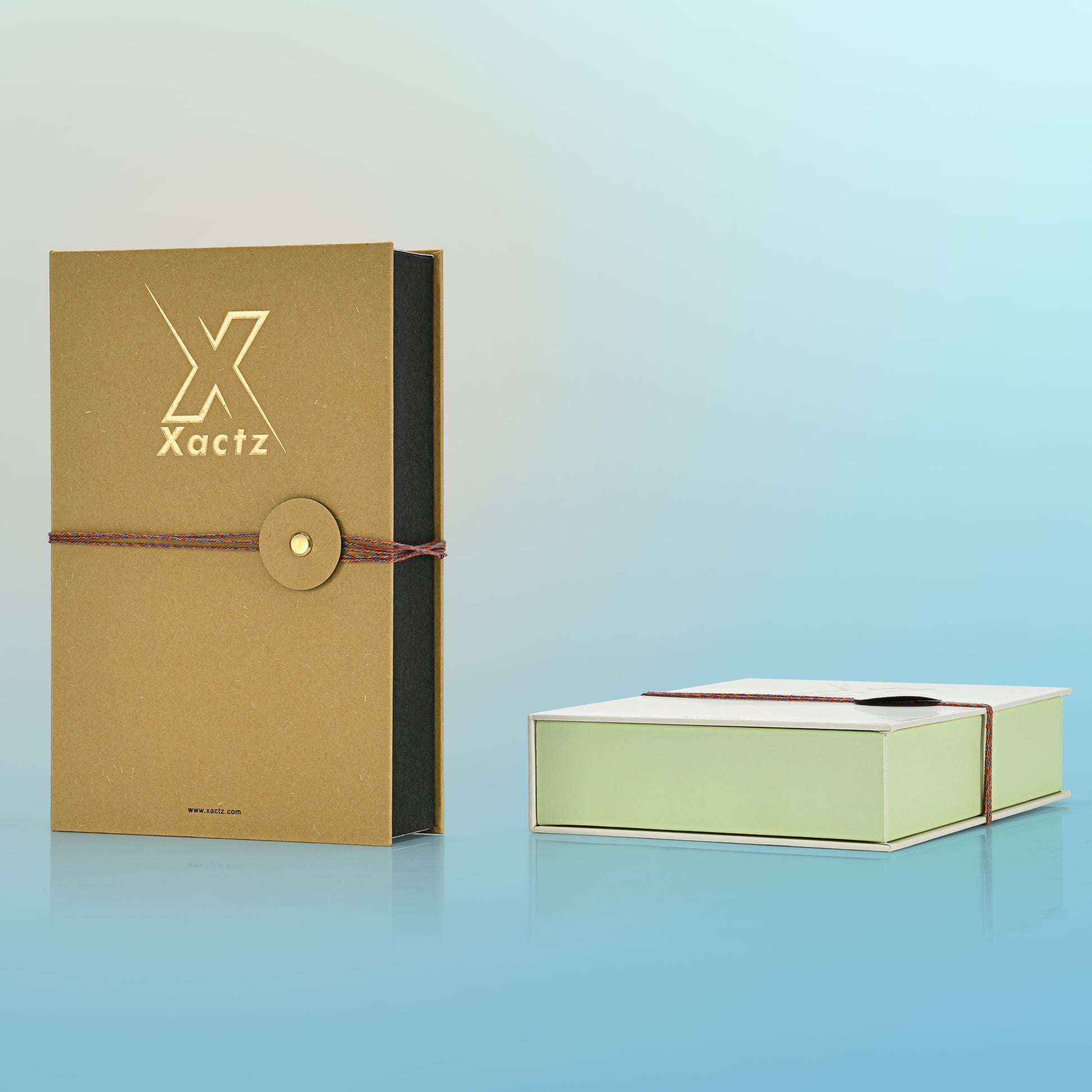 Product display showing a standing ochre yellow custom rigid book style perfume box next to a closed light grey and light green book style box lying flat on a reflective light blue background