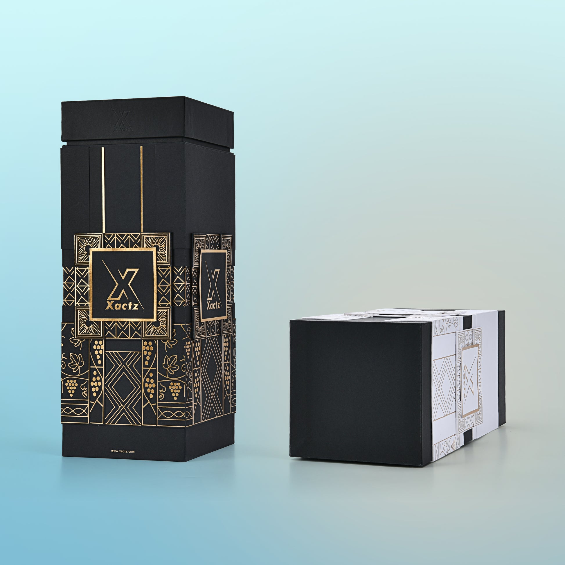 Lifestyle view of two custom rigid wine gift boxes showing the all black gold foil version standing upright with the matte black lid on and the white and black gold foil version placed horizontally beside it displaying the full collared base construction and the detailed gold foil grape vine and geometric illustration panels